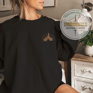 May include: Black sweatshirt with a gold castle design on the upper left chest. The design is a silhouette of a castle with a half-circle arch. The sweatshirt has the text "Embroidered Sweatshirt & Hoodie" in a circular design.