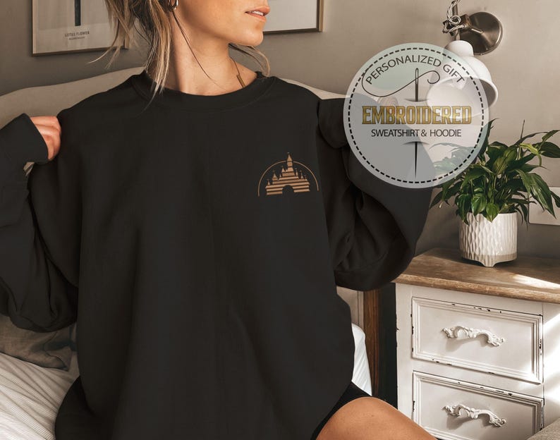 May include: Black sweatshirt with a gold castle design on the upper left chest. The design is a silhouette of a castle with a half-circle arch. The sweatshirt has the text "Embroidered Sweatshirt & Hoodie" in a circular design.