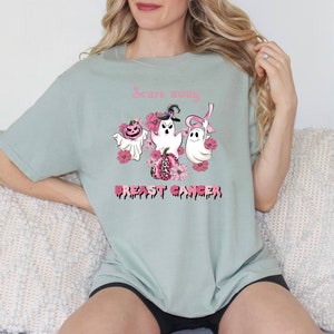 Pode incluir: Uma camiseta verde claro com uma fita rosa, fantasmas e uma ab&oacute;bora com o texto "Scare away Breast Cancer".