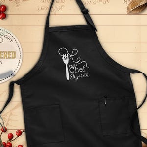 May include: Black apron with two front pockets and a pen holder. The apron features a white graphic of a fork and pasta, with the words "Pasta Chef Elizabeth" in a script font. The apron is a personalized gift.