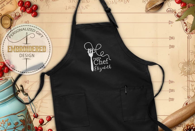 May include: Black apron with two front pockets and a pen holder. The apron features a white graphic of a fork and pasta, with the words "Pasta Chef Elizabeth" in a script font. The apron is a personalized gift.