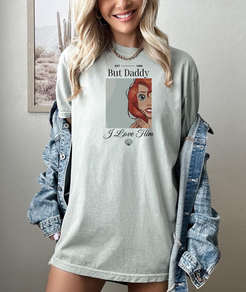 Może przedstawiać: Jasnozielona koszulka z grafiką z kresk&oacute;wki przedstawiającą rudowłosą postać, z napisem "But Daddy" i "I Love Him". Koszulka ma luźny kr&oacute;j i okrągły dekolt.