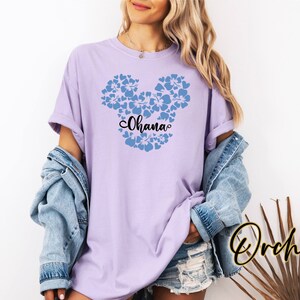 Puede incluir: Una camiseta lavanda con un dise&ntilde;o de Mickey Mouse floral azul y la palabra "Ohana" en texto negro.
