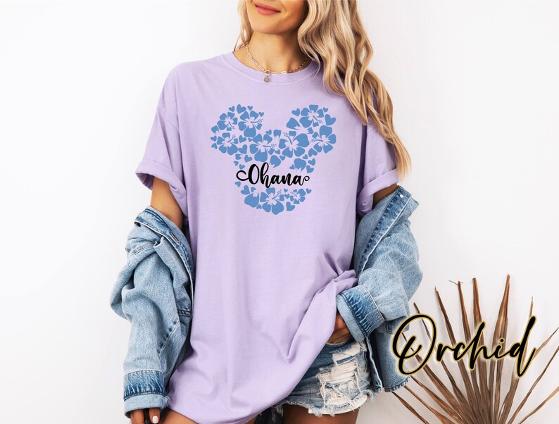 Puede incluir: Una camiseta lavanda con un dise&ntilde;o de Mickey Mouse floral azul y la palabra "Ohana" en texto negro.