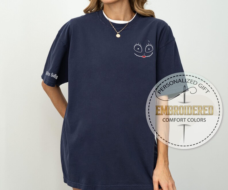 K&ouml;nnte beinhalten: Marineblaues, besticktes Comfort Colors T-Shirt mit einem Cartoon-Gesicht auf der Vorderseite und "His Sally" auf dem &Auml;rmel. Das Design hat einen wei&szlig;en Umriss mit schwarzen Augen und einem roten Mund. Das Shirt ist ein personalisiertes Geschenk.