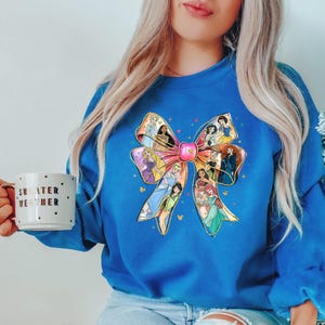 May include: A royal blue sweatshirt features a large bow design with various princess illustrations. The bow is pink with gold accents and the princesses are in their signature outfits. A white mug with the words "SWEATER WEATHER" is held in the hand.