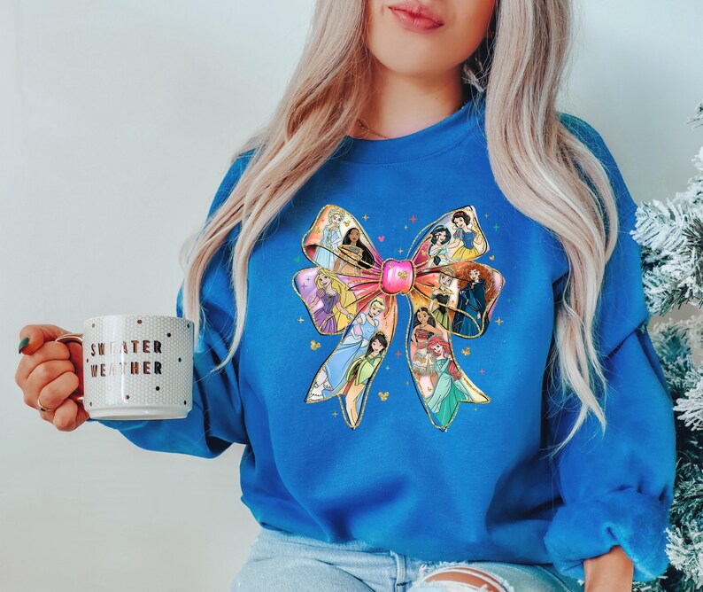 May include: A royal blue sweatshirt features a large bow design with various princess illustrations. The bow is pink with gold accents and the princesses are in their signature outfits. A white mug with the words "SWEATER WEATHER" is held in the hand.