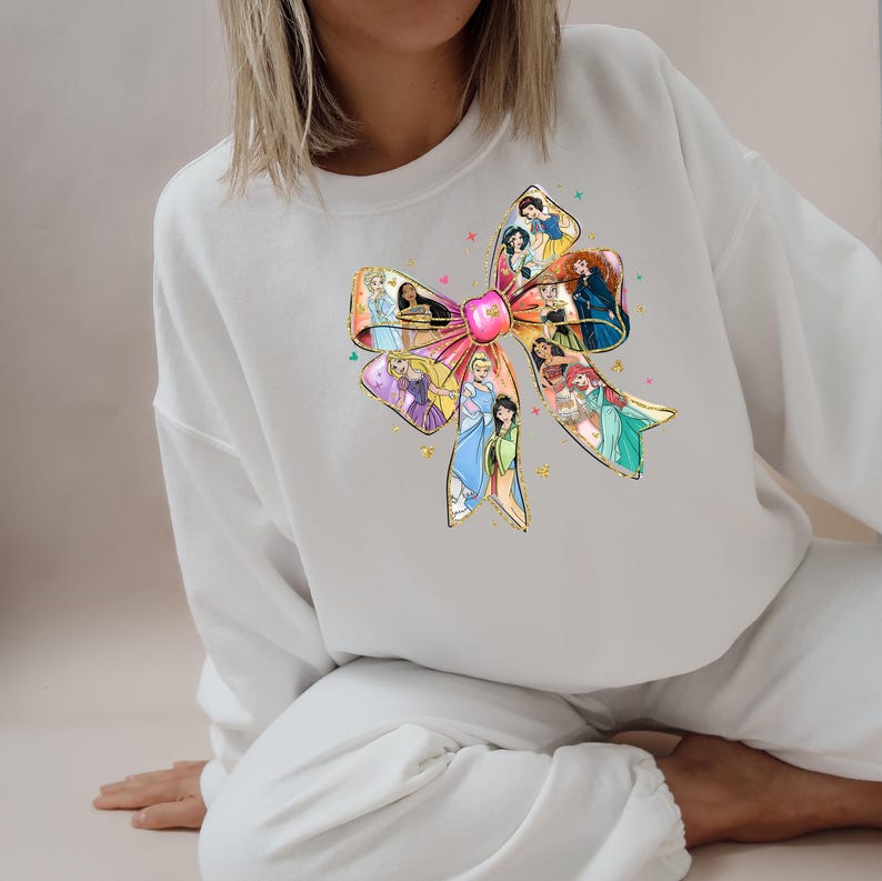 May include: White sweatshirt featuring a large, colorful bow design. The bow is adorned with illustrations of various princesses. The design includes gold accents and a pink center, creating a whimsical and eye-catching aesthetic.