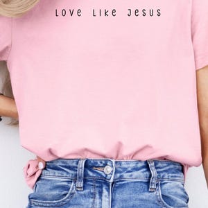 May include: A light pink t-shirt with the text "Love Like Jesus" in black lettering. The shirt is paired with blue denim jeans. The overall style is casual and comfortable.