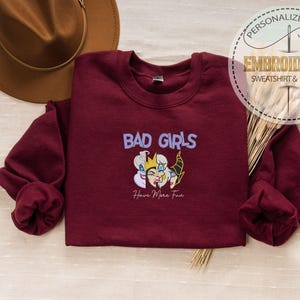 Puede incluir: Una sudadera granate con las palabras "BAD GIRLS" en morado y una ilustraci&oacute;n de tres personajes villanos. El texto "Have More Fun" est&aacute; debajo de la ilustraci&oacute;n. Un sombrero marr&oacute;n es visible en el fondo.
