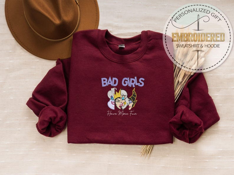 Puede incluir: Una sudadera granate con las palabras "BAD GIRLS" en morado y una ilustraci&oacute;n de tres personajes villanos. El texto "Have More Fun" est&aacute; debajo de la ilustraci&oacute;n. Un sombrero marr&oacute;n es visible en el fondo.
