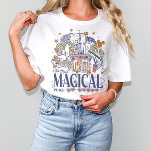 May include: White t-shirt featuring a colorful graphic design with the text "The Most Magical Place on Earth." The design includes illustrations of a castle, teacups, and other theme park elements. The shirt is a classic crew neck style.