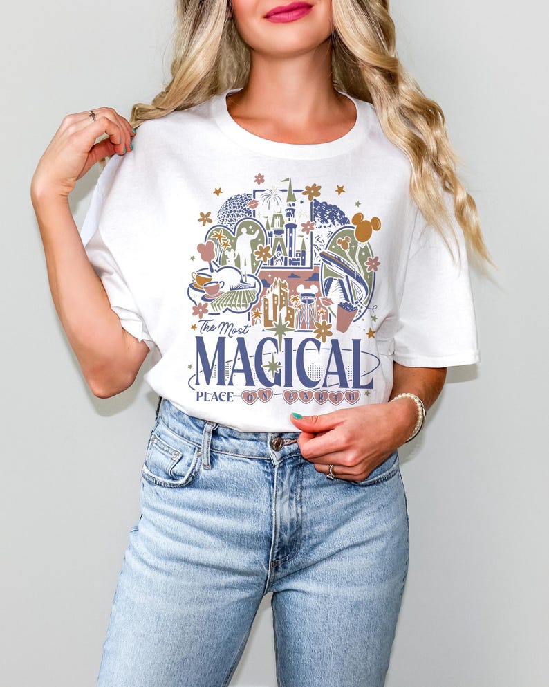 May include: White t-shirt featuring a colorful graphic design with the text "The Most Magical Place on Earth." The design includes illustrations of a castle, teacups, and other theme park elements. The shirt is a classic crew neck style.