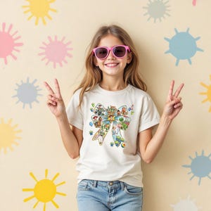 May include: A light-colored t-shirt featuring a colorful bow design with various cartoon characters. The girl is wearing pink sunglasses and blue jeans. The background is a light beige color with colorful sun and starburst shapes.