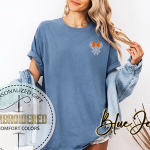 Pu&ograve; includere: Una maglietta blu con un fiocco arancione ricamato e un disegno a ragnatela. La maglietta ha le maniche arrotolate e le parole "Personalized Gift", "Embroidered" e "Comfort Colors" sono stampate su una grafica circolare. Le parole "Blue Jean" sono scritte in corsivo.