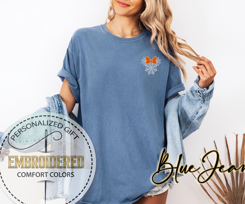 Pu&ograve; includere: Una maglietta blu con un fiocco arancione ricamato e un disegno a ragnatela. La maglietta ha le maniche arrotolate e le parole "Personalized Gift", "Embroidered" e "Comfort Colors" sono stampate su una grafica circolare. Le parole "Blue Jean" sono scritte in corsivo.