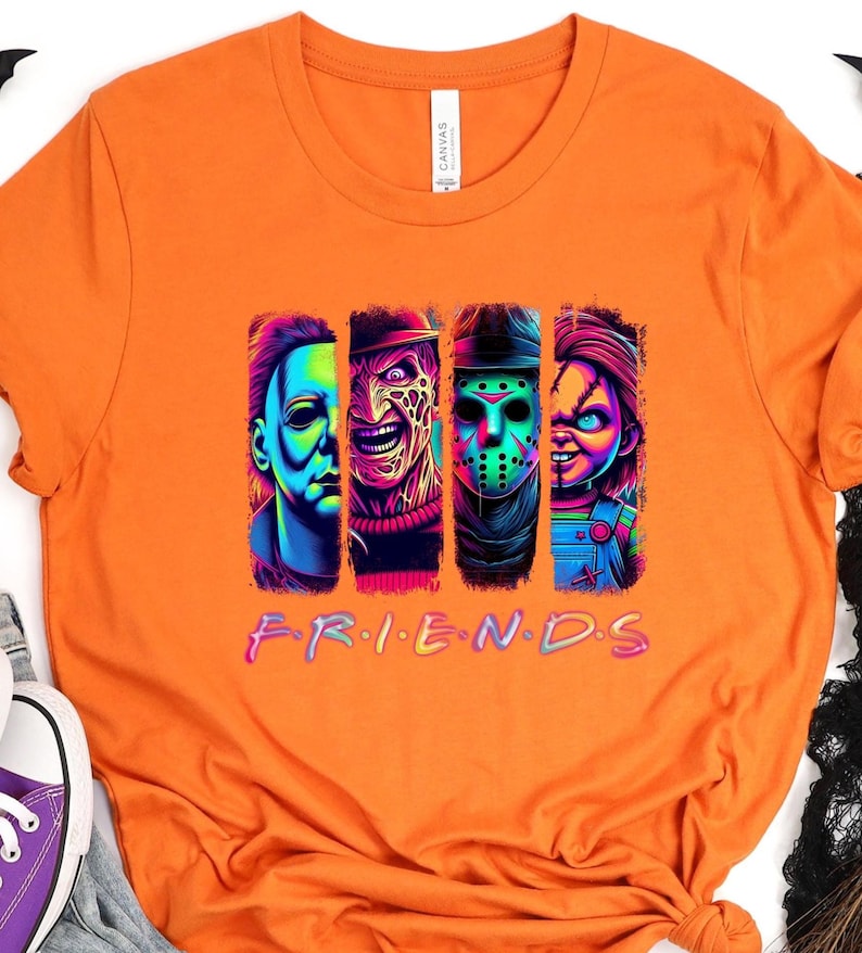 May include: Orange t-shirt featuring a graphic of four horror movie characters in vertical panels. The word "FRIENDS" is written in a colorful, stylized font below the characters. The shirt is made of a soft material.