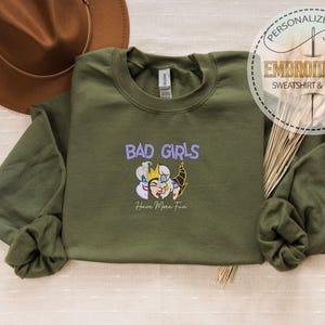 Puede incluir: Sudadera verde oliva con el texto bordado "BAD GIRLS" sobre un gr&aacute;fico de dibujos animados. El gr&aacute;fico presenta tres personajes de dibujos animados. Debajo del gr&aacute;fico est&aacute; el texto "Have More Fun". Un sombrero marr&oacute;n est&aacute; en el fondo.