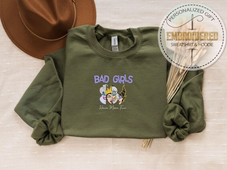 Puede incluir: Sudadera verde oliva con el texto bordado "BAD GIRLS" sobre un gr&aacute;fico de dibujos animados. El gr&aacute;fico presenta tres personajes de dibujos animados. Debajo del gr&aacute;fico est&aacute; el texto "Have More Fun". Un sombrero marr&oacute;n est&aacute; en el fondo.