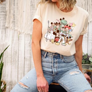 May include: Cream-colored t-shirt featuring a colorful cartoon graphic of various Disney characters, including Mickey Mouse, Woody, and Buzz Lightyear. The design includes balloons and other playful elements.