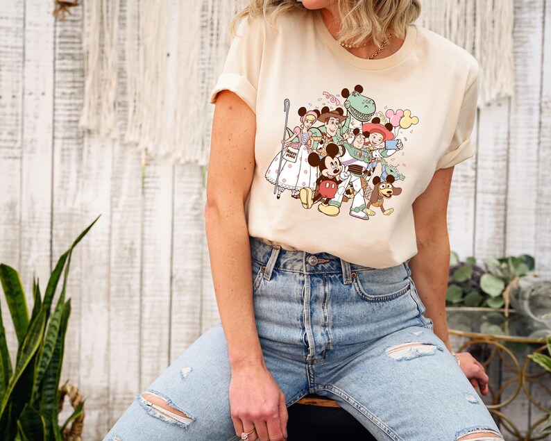 May include: Cream-colored t-shirt featuring a colorful cartoon graphic of various Disney characters, including Mickey Mouse, Woody, and Buzz Lightyear. The design includes balloons and other playful elements.