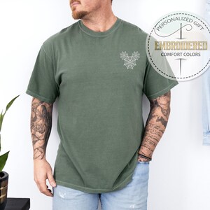Pu&ograve; includere: T-shirt verde oliva con un disegno a ragnatela ricamato bianco sul petto. La maglietta ha maniche corte e girocollo. Il testo "PERSONALIZED GIFT EMBROIDERED COMFORT COLORS" &egrave; stampato su un'etichetta circolare.