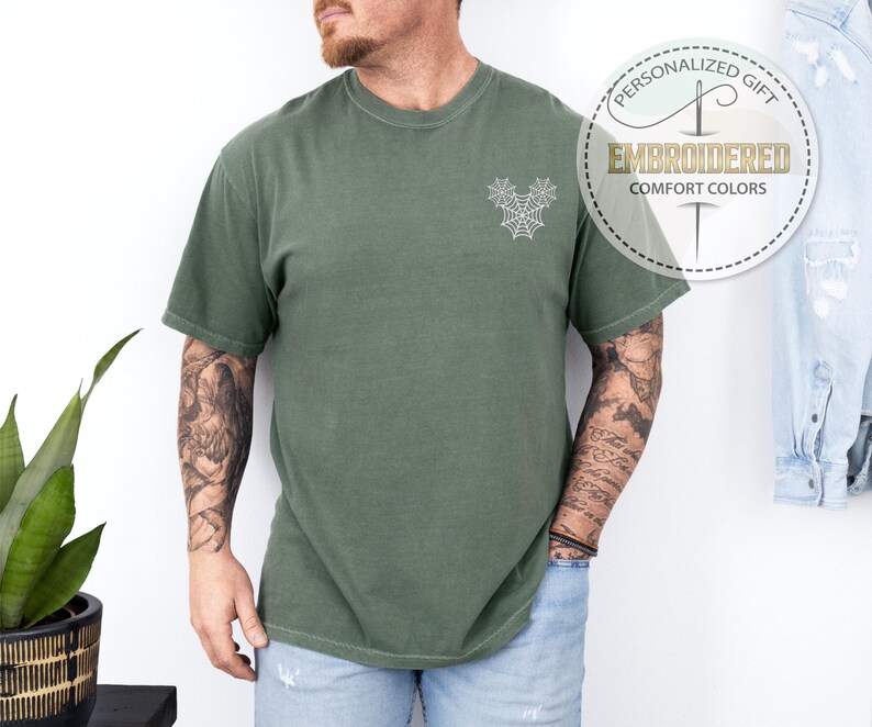 Pu&ograve; includere: T-shirt verde oliva con un disegno a ragnatela ricamato bianco sul petto. La maglietta ha maniche corte e girocollo. Il testo "PERSONALIZED GIFT EMBROIDERED COMFORT COLORS" &egrave; stampato su un'etichetta circolare.