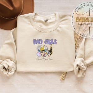 Puede incluir: Sudadera color crema con el texto "BAD GIRLS" en morado y un gr&aacute;fico de tres villanos de dibujos animados. Debajo del gr&aacute;fico est&aacute; el texto "Have More Fun". Una etiqueta de regalo personalizada est&aacute; en la esquina superior derecha.