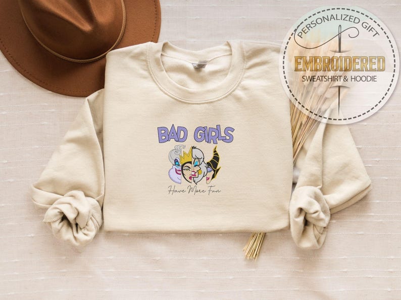 Puede incluir: Sudadera color crema con el texto "BAD GIRLS" en morado y un gr&aacute;fico de tres villanos de dibujos animados. Debajo del gr&aacute;fico est&aacute; el texto "Have More Fun". Una etiqueta de regalo personalizada est&aacute; en la esquina superior derecha.