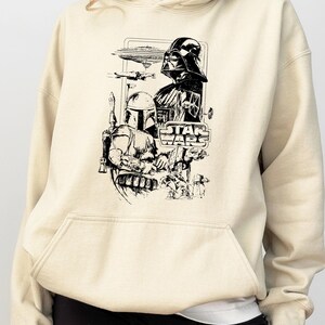 May include: Cream-colored hoodie featuring a black and white graphic of Star Wars characters, including Darth Vader and Boba Fett. The Star Wars logo is also visible. The hoodie has a front pocket and a drawstring hood.