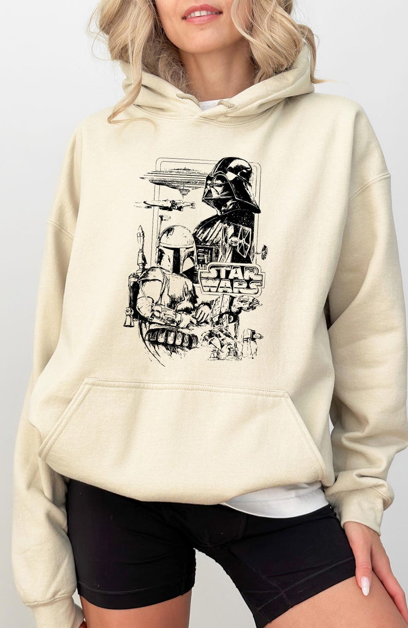 May include: Cream-colored hoodie featuring a black and white graphic of Star Wars characters, including Darth Vader and Boba Fett. The Star Wars logo is also visible. The hoodie has a front pocket and a drawstring hood.
