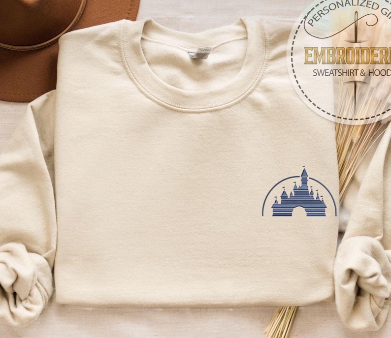 May include: Cream-colored sweatshirt with a blue castle graphic on the front. The graphic is a stylized illustration of a castle within a half-circle. The sweatshirt has a crew neck and long sleeves.