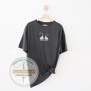Pu&ograve; includere: T-shirt grigio antracite con un disegno ricamato di due oche bianche con becchi e zampe arancioni. Il testo "GOOSE BUMPS" &egrave; sotto. La maglietta &egrave; appesa a una gruccia di legno su sfondo bianco.