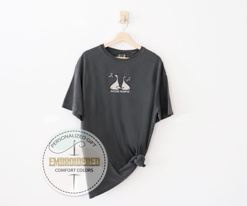 Pu&ograve; includere: T-shirt grigio antracite con un disegno ricamato di due oche bianche con becchi e zampe arancioni. Il testo "GOOSE BUMPS" &egrave; sotto. La maglietta &egrave; appesa a una gruccia di legno su sfondo bianco.