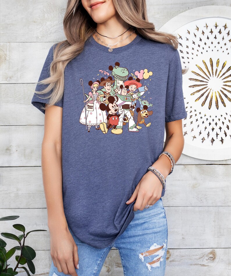 May include: A navy blue t-shirt featuring a colorful graphic of Disney and Pixar characters, including Mickey Mouse, Buzz Lightyear, and others. The design is a cartoon-style illustration. The shirt has short sleeves and a crew neck.