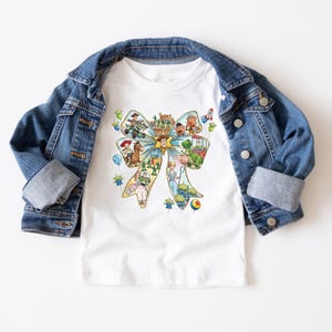 May include: White t-shirt featuring a colorful bow design with various cartoon characters. The shirt is paired with a blue denim jacket. The bow design includes characters from a popular animated movie.