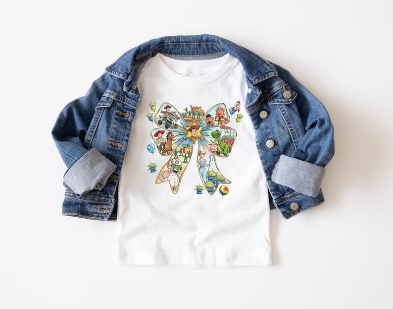 May include: White t-shirt featuring a colorful bow design with various cartoon characters. The shirt is paired with a blue denim jacket. The bow design includes characters from a popular animated movie.
