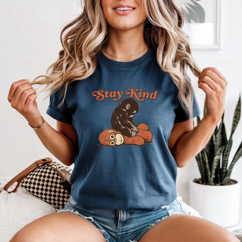 May include: A navy blue t-shirt featuring the text "Stay Kind" in orange script above a graphic of a monkey hugging a teddy bear. The monkey is black and the teddy bear is orange. The shirt has short sleeves.