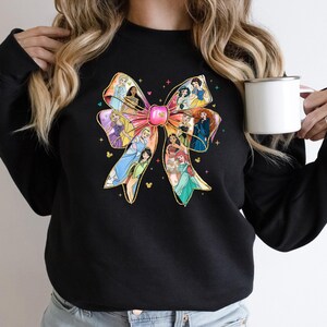 May include: Black sweatshirt featuring a colorful bow design with various princess illustrations. The bow has a pink center and gold accents, with a Mickey Mouse head at the bottom of the bow. The sweatshirt is a crew neck style.