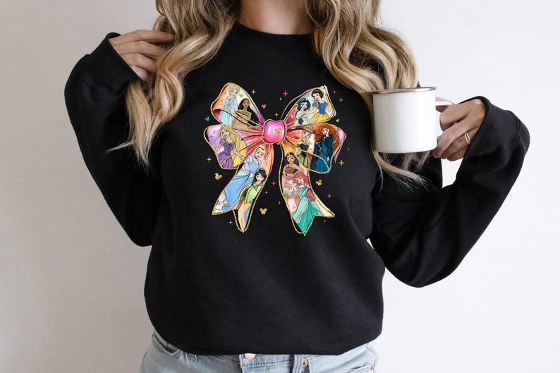May include: Black sweatshirt featuring a colorful bow design with various princess illustrations. The bow has a pink center and gold accents, with a Mickey Mouse head at the bottom of the bow. The sweatshirt is a crew neck style.