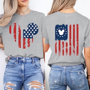 May include: A gray t-shirt featuring a distressed American flag design with a Mickey Mouse silhouette. The front design is a Mickey Mouse head shape with red stripes and a blue circle filled with white stars. The back has a flag design.