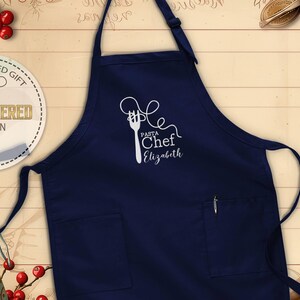 May include: A navy blue apron with a white embroidered design of a fork and pasta, with the words "Pasta Chef Elizabeth." The apron has two pockets and a pen holder. The apron is a personalized gift.