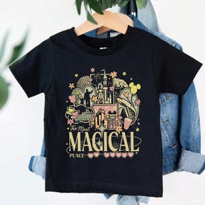 May include: Black t-shirt with a colorful graphic featuring a castle, flowers, and Mickey Mouse ears. The text "The Most Magical Peace" is displayed in a decorative font. The shirt is hanging on a wooden hanger.