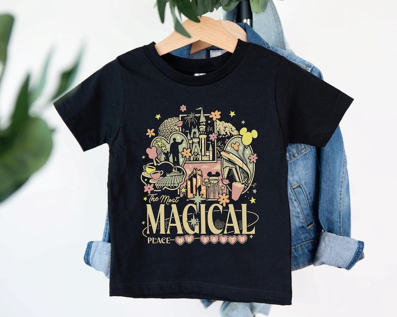 May include: Black t-shirt with a colorful graphic featuring a castle, flowers, and Mickey Mouse ears. The text "The Most Magical Peace" is displayed in a decorative font. The shirt is hanging on a wooden hanger.