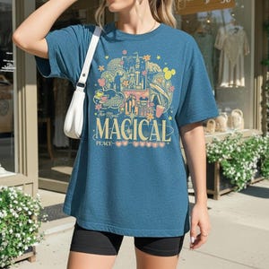 May include: A teal blue t-shirt with a colorful graphic design featuring a castle, Mickey Mouse ears, and the text "The Most Magical Place". The shirt is a relaxed fit.