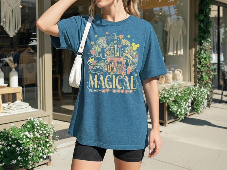 May include: A teal blue t-shirt with a colorful graphic design featuring a castle, Mickey Mouse ears, and the text "The Most Magical Place". The shirt is a relaxed fit.