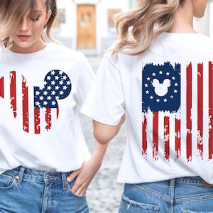 May include: White t-shirts with a distressed American flag design featuring a Mickey Mouse silhouette. The front design is a Mickey Mouse head with red stripes and a blue circle filled with white stars. The back design is a distressed American flag with a Mickey Mouse head in the blue field.