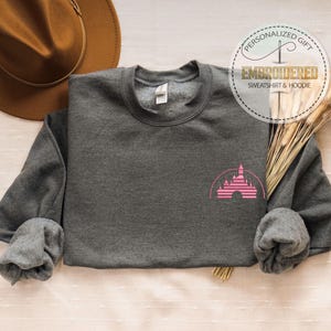 May include: A dark gray sweatshirt with a pink castle graphic on the front. The sweatshirt has long sleeves and a crew neck. The image also includes a brown hat, dried wheat, and a circular logo that says "Embroidered Sweatshirt & Hoodie".