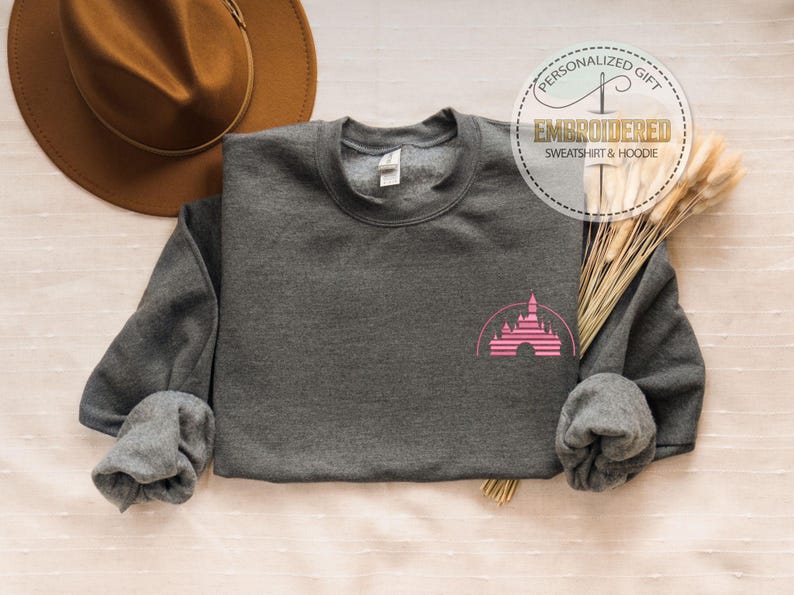 May include: A dark gray sweatshirt with a pink castle graphic on the front. The sweatshirt has long sleeves and a crew neck. The image also includes a brown hat, dried wheat, and a circular logo that says "Embroidered Sweatshirt & Hoodie".