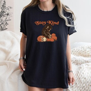 May include: Black t-shirt with the text "Stay Kind" in orange lettering. The shirt features a graphic of a brown ape hugging a teddy bear. The shirt is a relaxed fit.